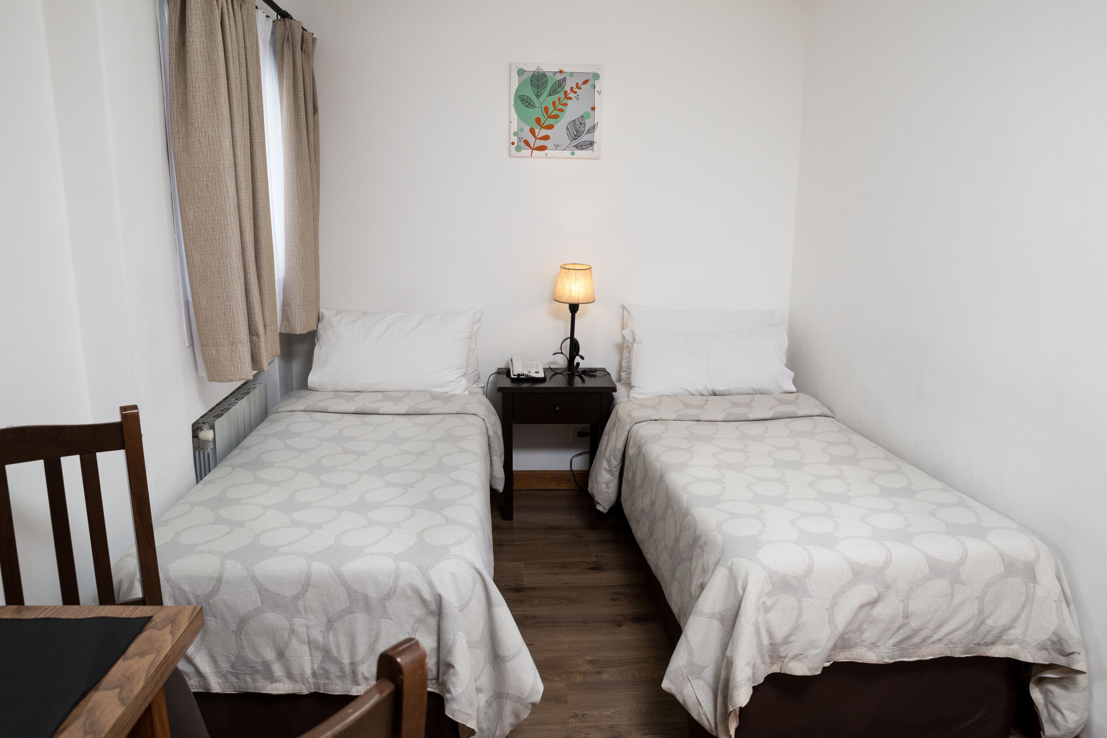 Una habitación con dos camas individuales, una mesita de noche con lámpara, una silla y una ventana con cortinas.