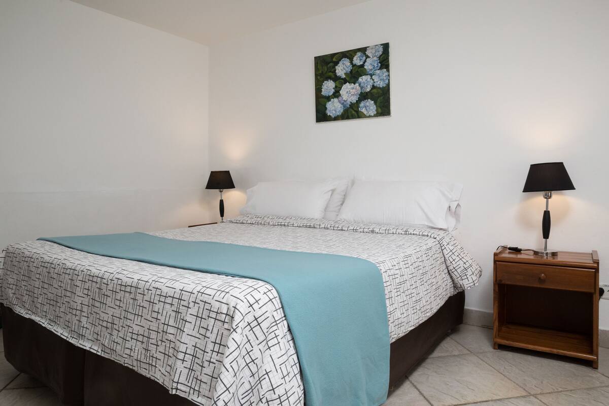 Ein ordentlich bezogenes Bett mit einem gemusterten Überwurf, zwei Nachttischlampen, einem kleinen Holfnachtkästchen und einem an der Wand hängenden Blumenbild.