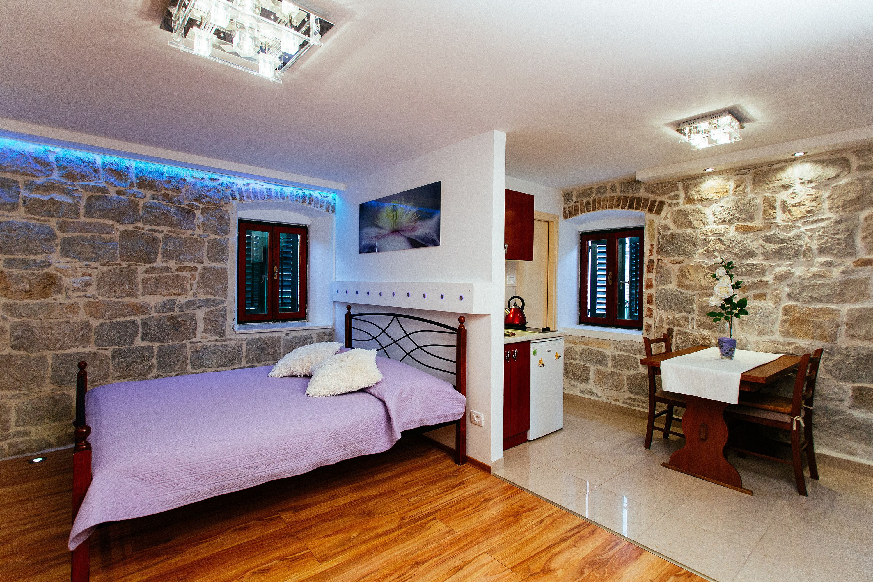 A bedroom with a bed, a small table, and a chair, a kitchenette, and stone walls.