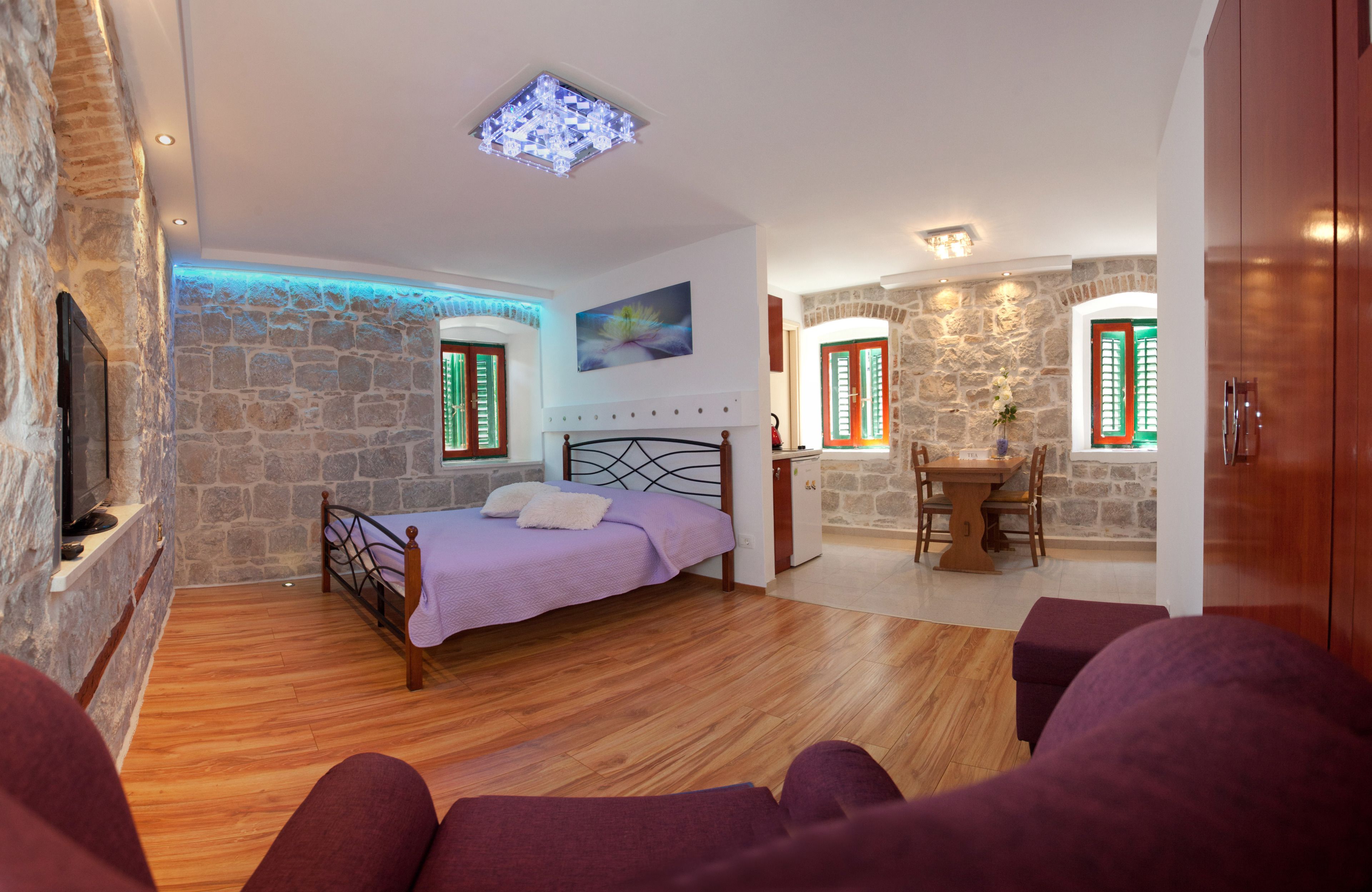 A bedroom with a bed, a TV, stone walls, a dining area, and a kitchen.