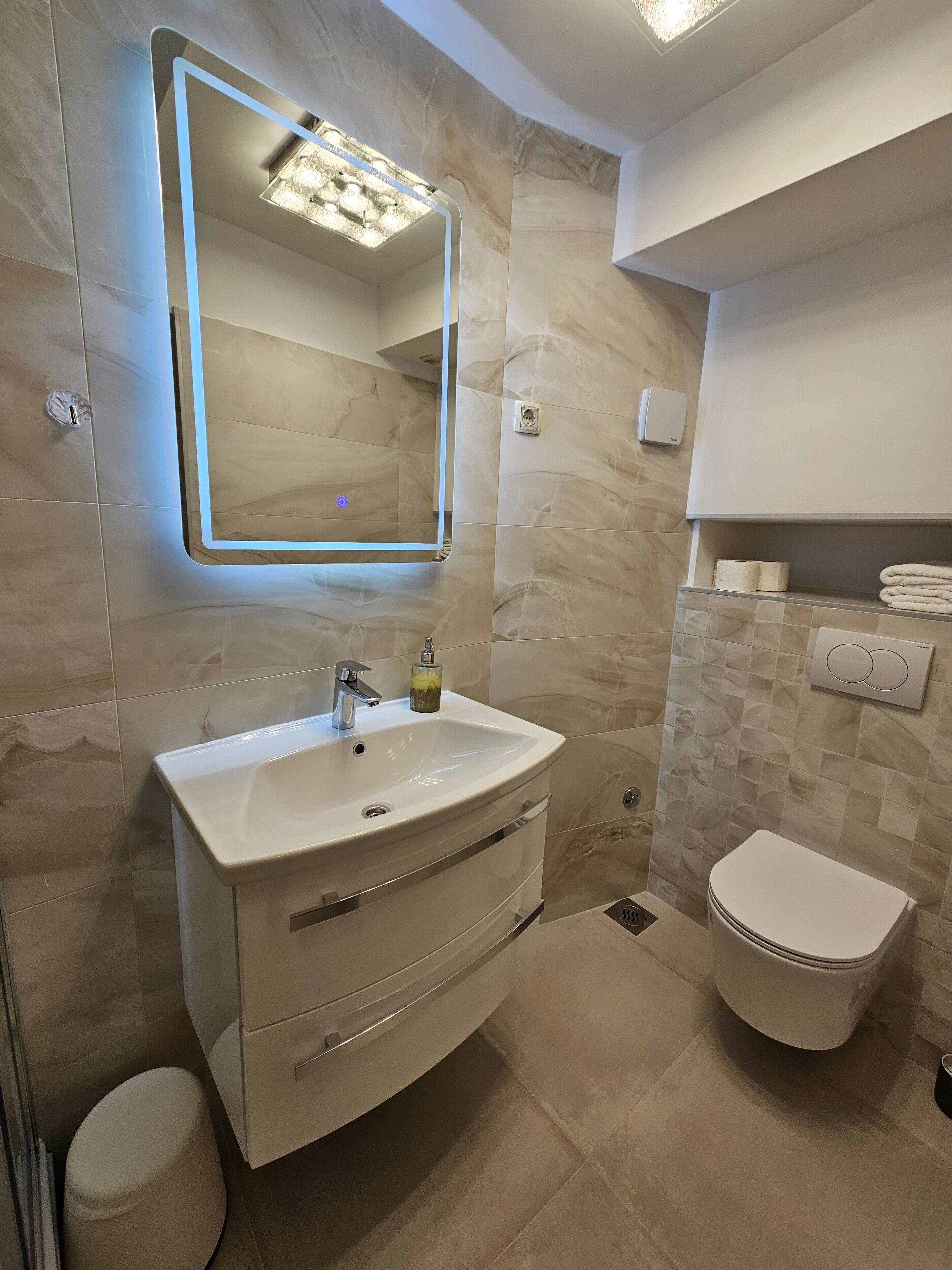 A modern bathroom with a white vanity, a mirror with under-cabinet lighting, a toilet, and a towel rack.