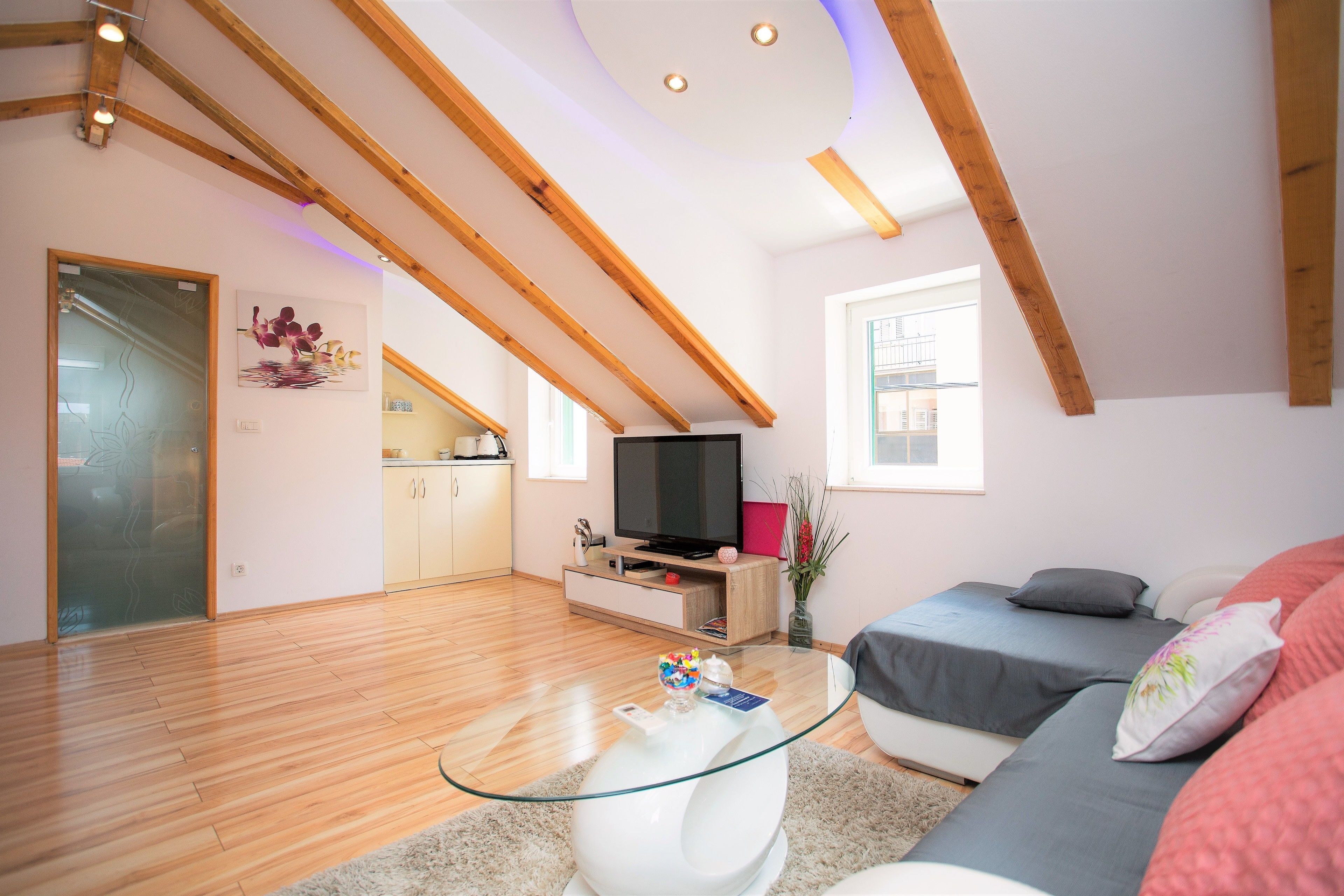 Ein modernes Wohnzimmer mit Glastisch, grauem Sofa, Holzbalken an der Decke, einem Fernseher und einer Küche im Hintergrund.