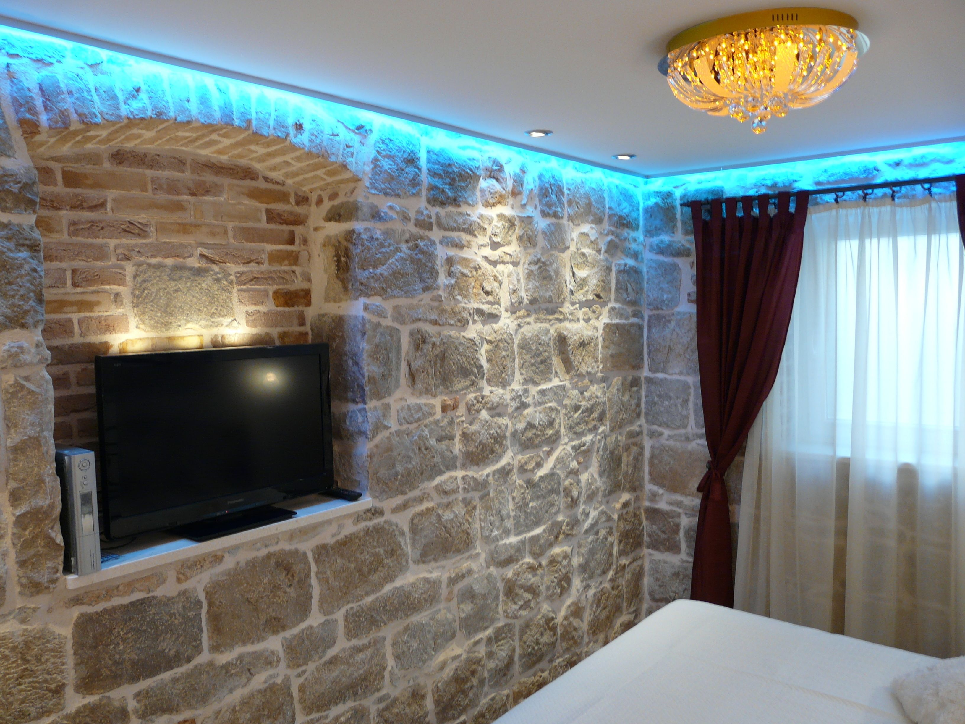Una habitación con pared de piedra, televisor de pantalla plana montado en ella, cama y candelabro.