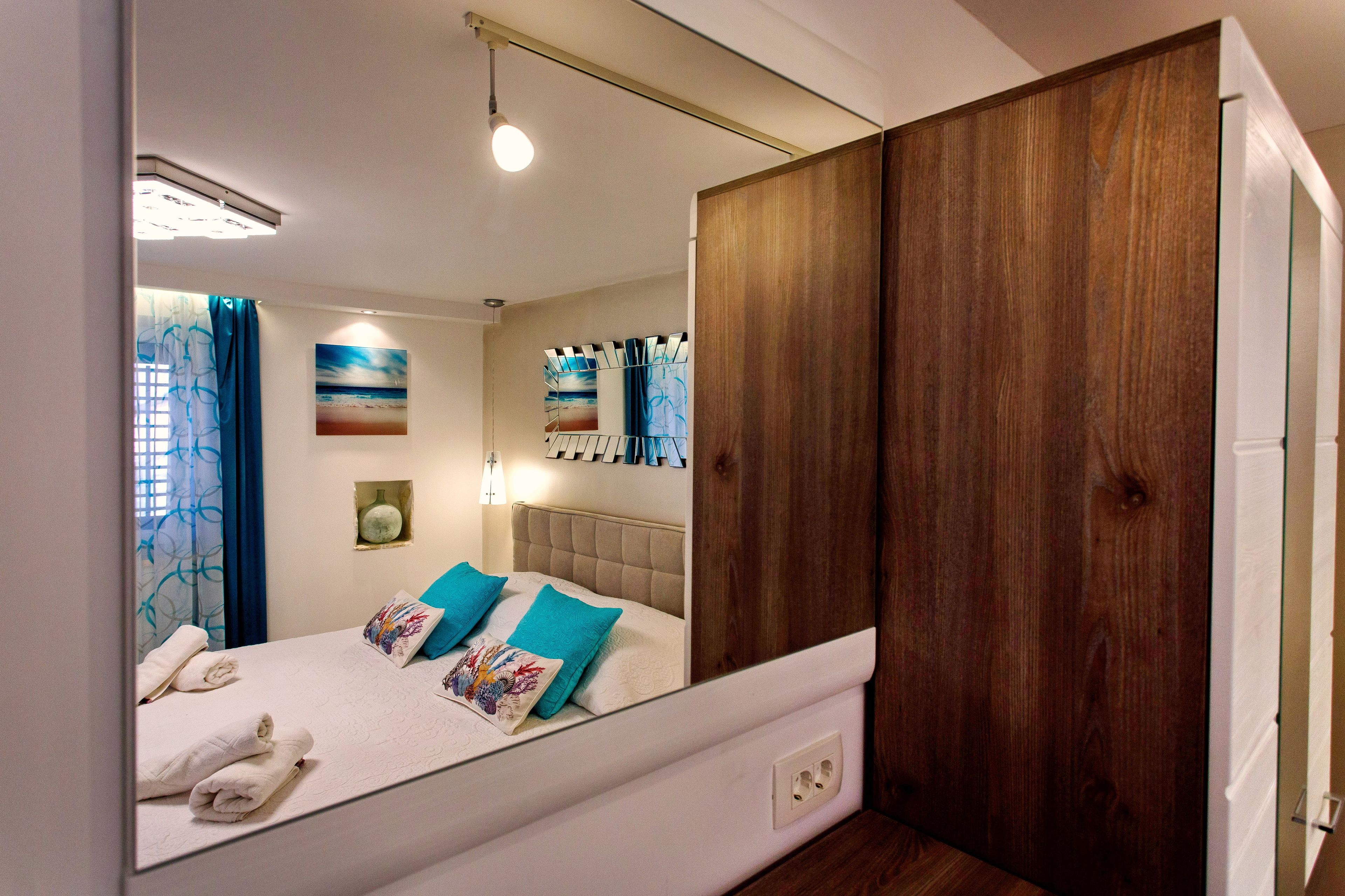 Un dormitorio con un espejo grande, una cama con ropa de cama azul y blanca, un armario de madera y obras de arte en las paredes.