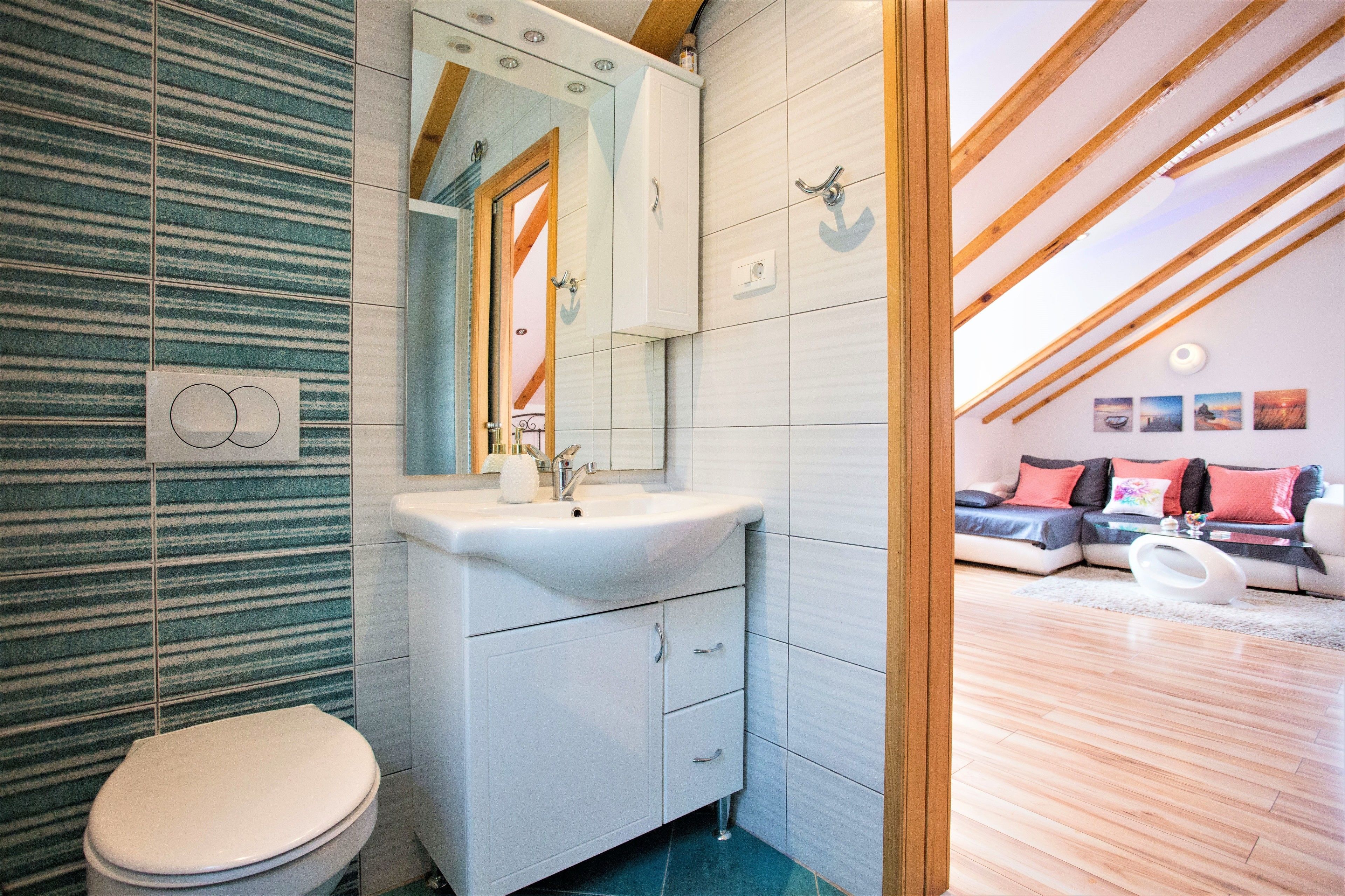 Une salle de bain moderne avec un lavabo blanc, un miroir et des toilettes. On y trouve une poutre en bois au plafond et, au fond, un espace de vie avec un canapé et des coussins.