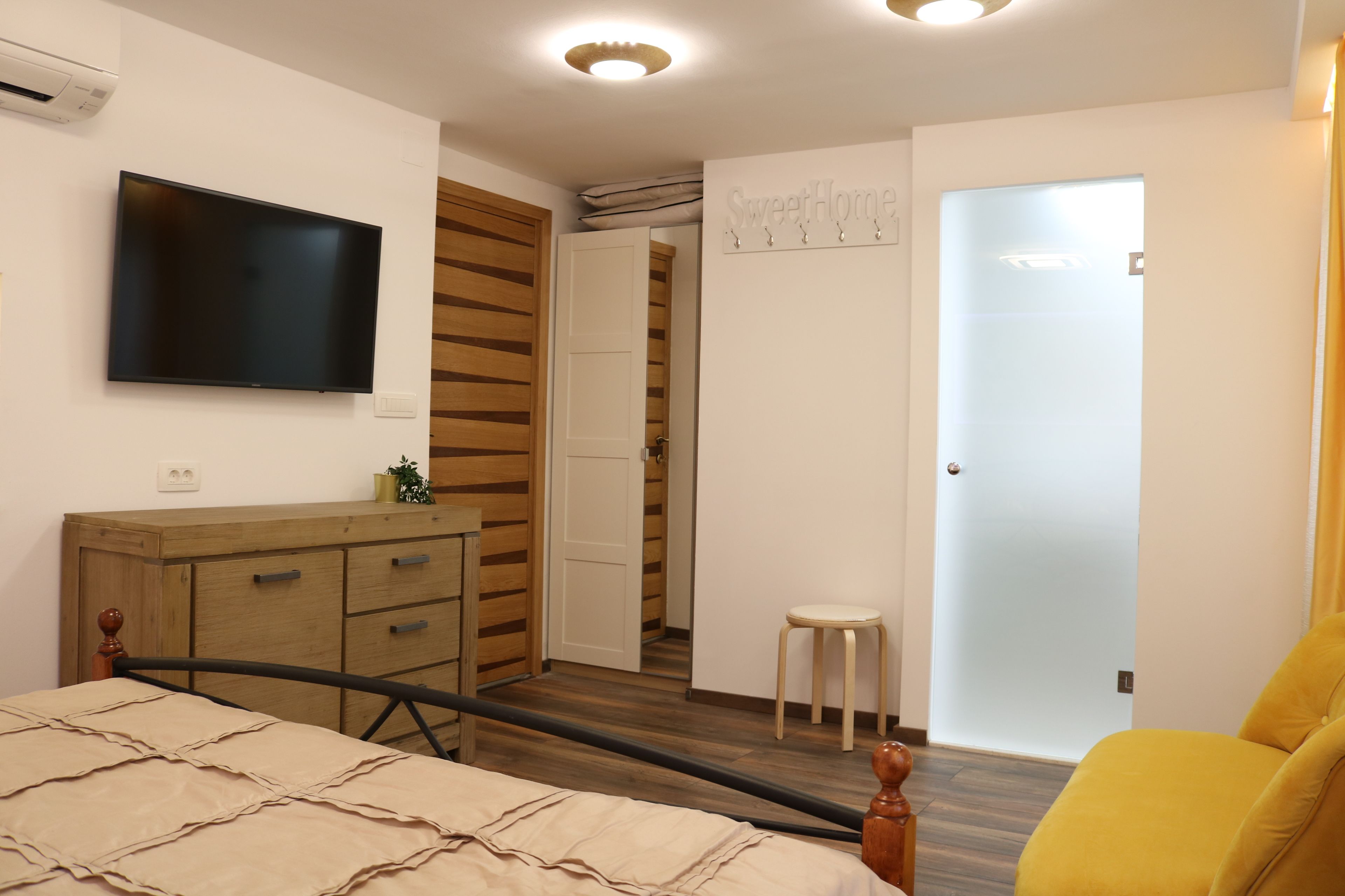 A bedroom with a bed, a wooden dresser, a television, a small stool, and a door leading to another room.