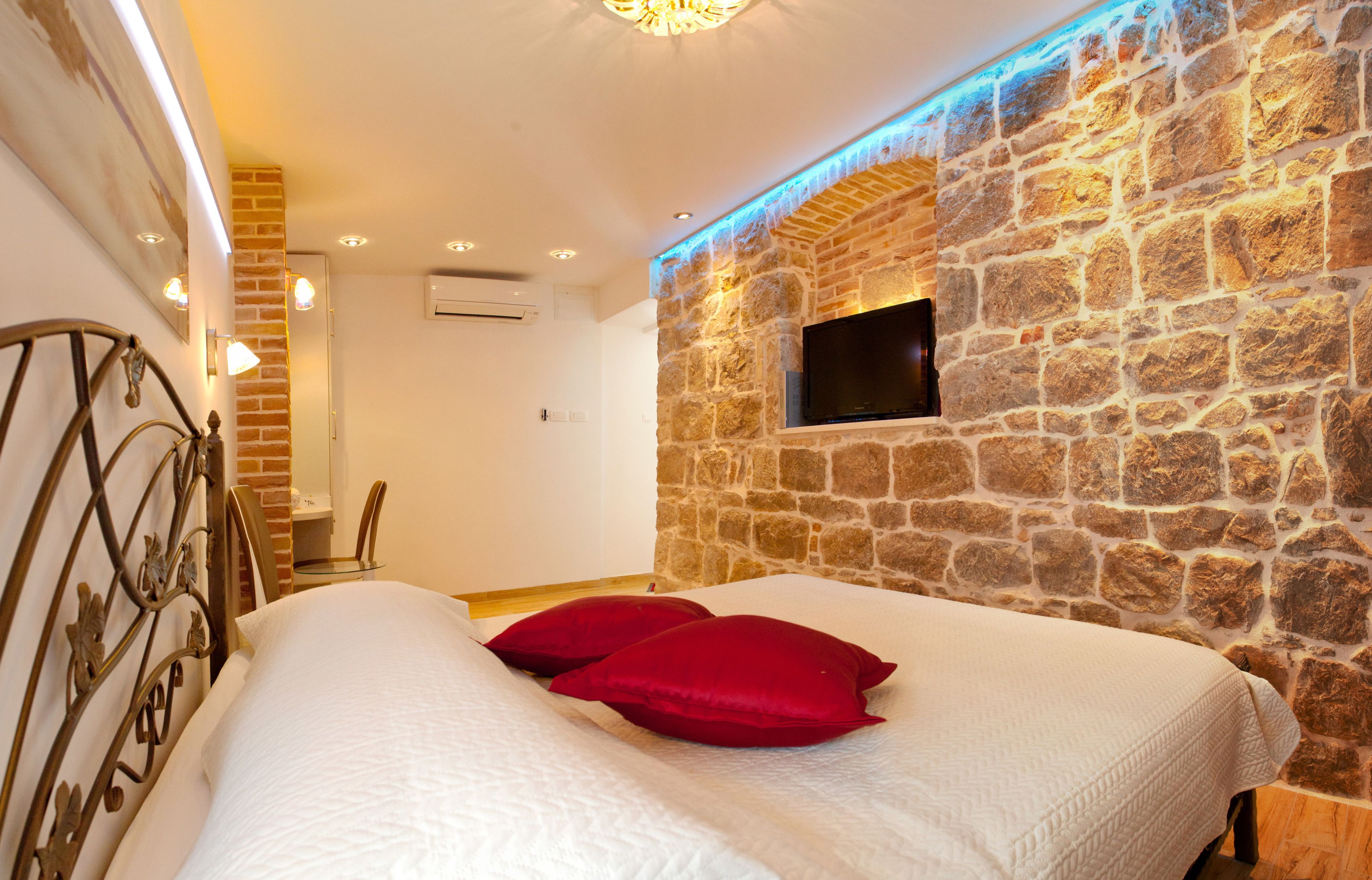 Un dormitorio con pared de piedra, televisor de pantalla plana, cama con ropa de cama blanca y almohadas rojas, y cabecero de metal.