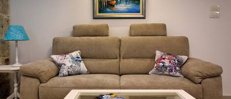 A living room with a beige sofa, two patterned cushions, a glass coffee table with magazines, a lamp, and a framed painting on the wall.
