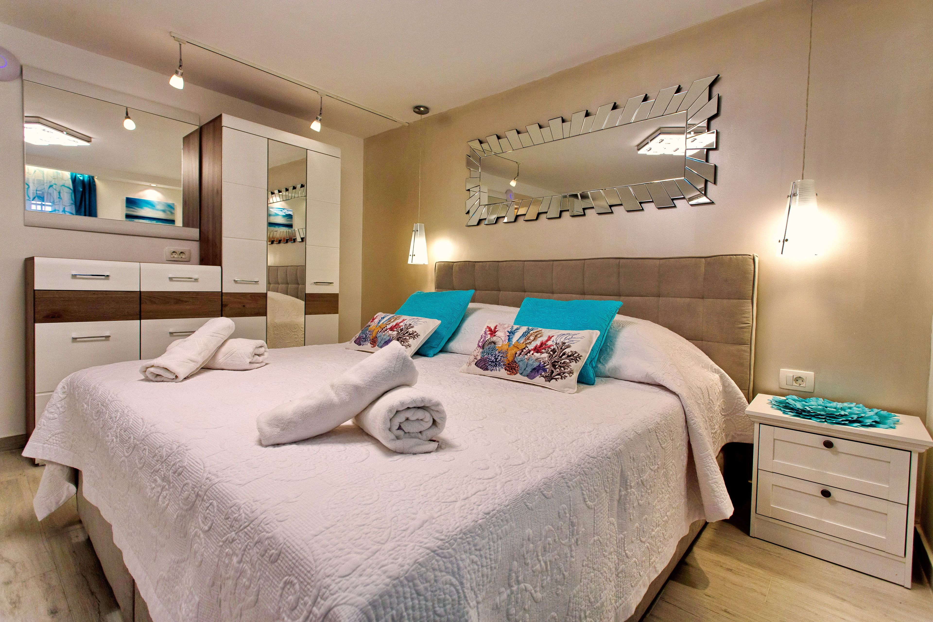 A neatly made bed with a white bedspread and two decorative pillows, a bedside table with a lamp, and a large mirror on the wall.