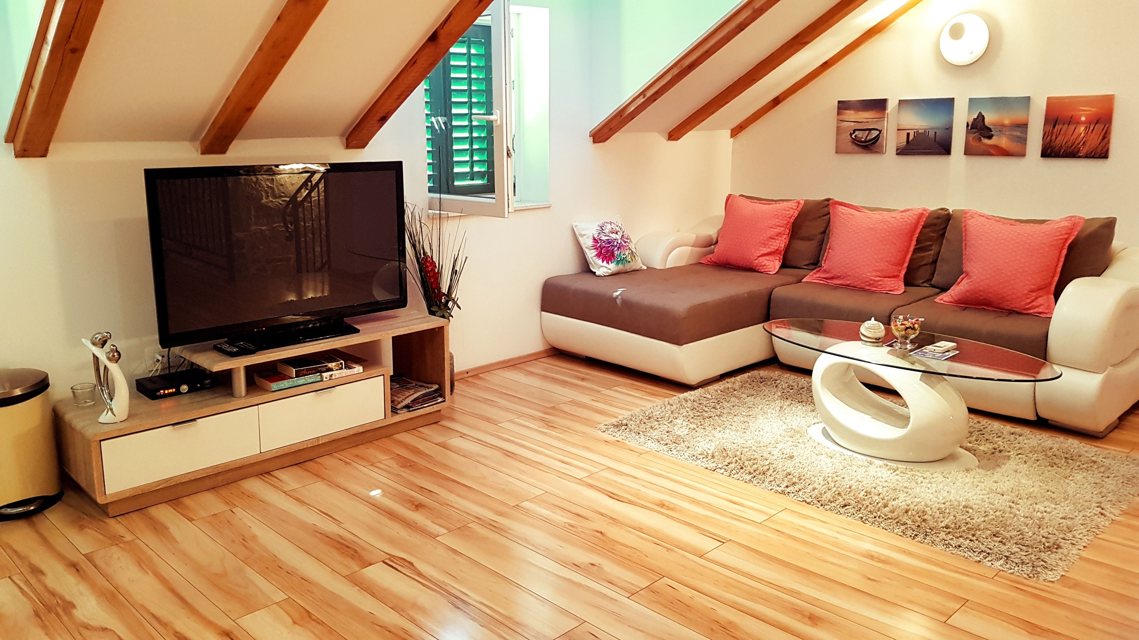 A modern living room with a wooden floor, a flat-screen TV, a sofa with red cushions, and a glass coffee table.