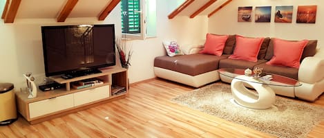 A modern living room with a wooden floor, a flat-screen TV, a sofa with red cushions, and a glass coffee table.