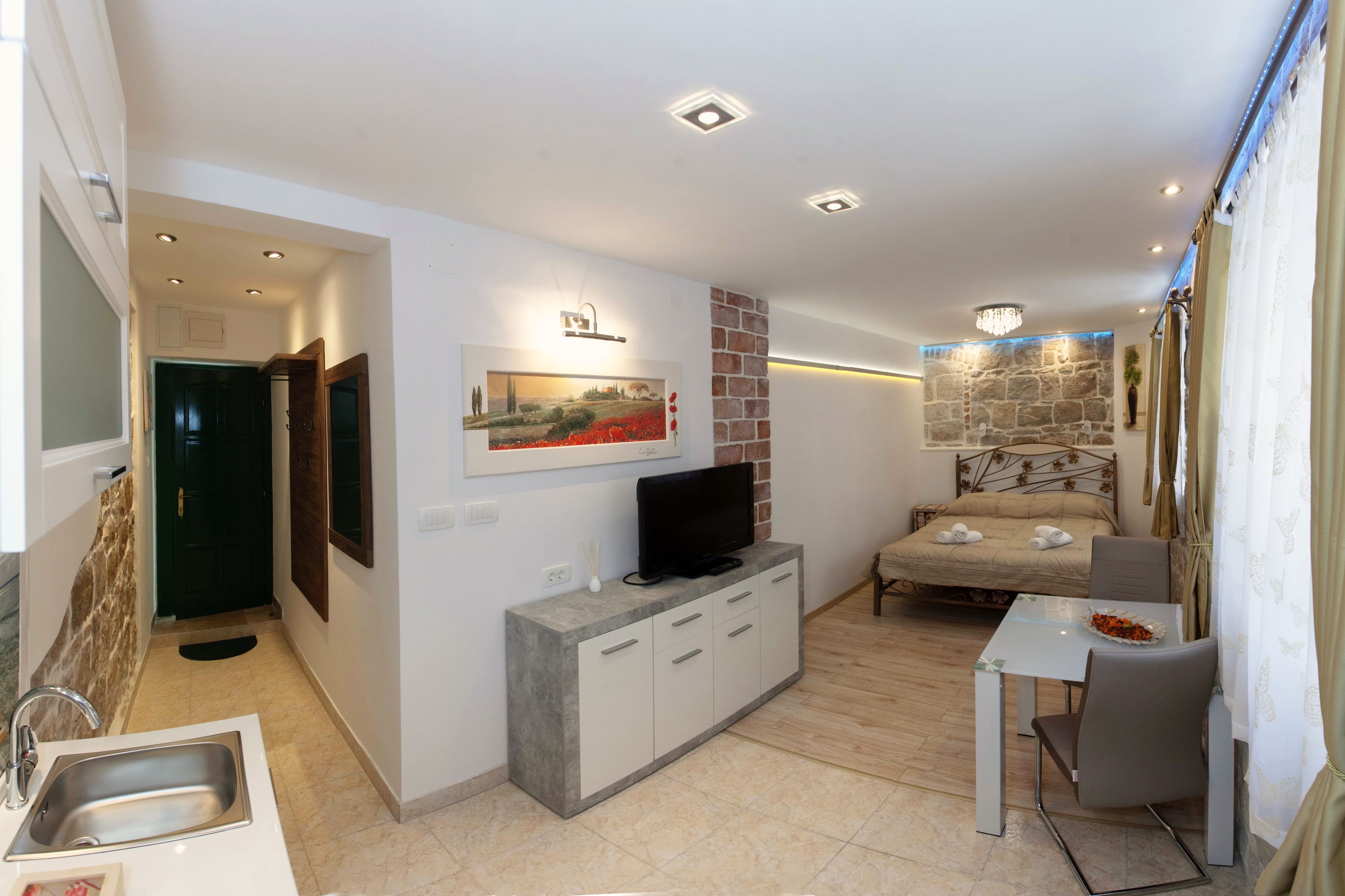 A modern living room with a flat-screen TV, a stone accent wall, a dining area with a table and chairs, and a small kitchen with a sink and cabinets.