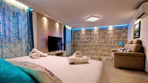 A modern hotel room with a large bed, a flat-screen TV, a stone accent wall, a seating area with a sofa and armchairs, and a small table with a lamp. - House Sandra (Split)