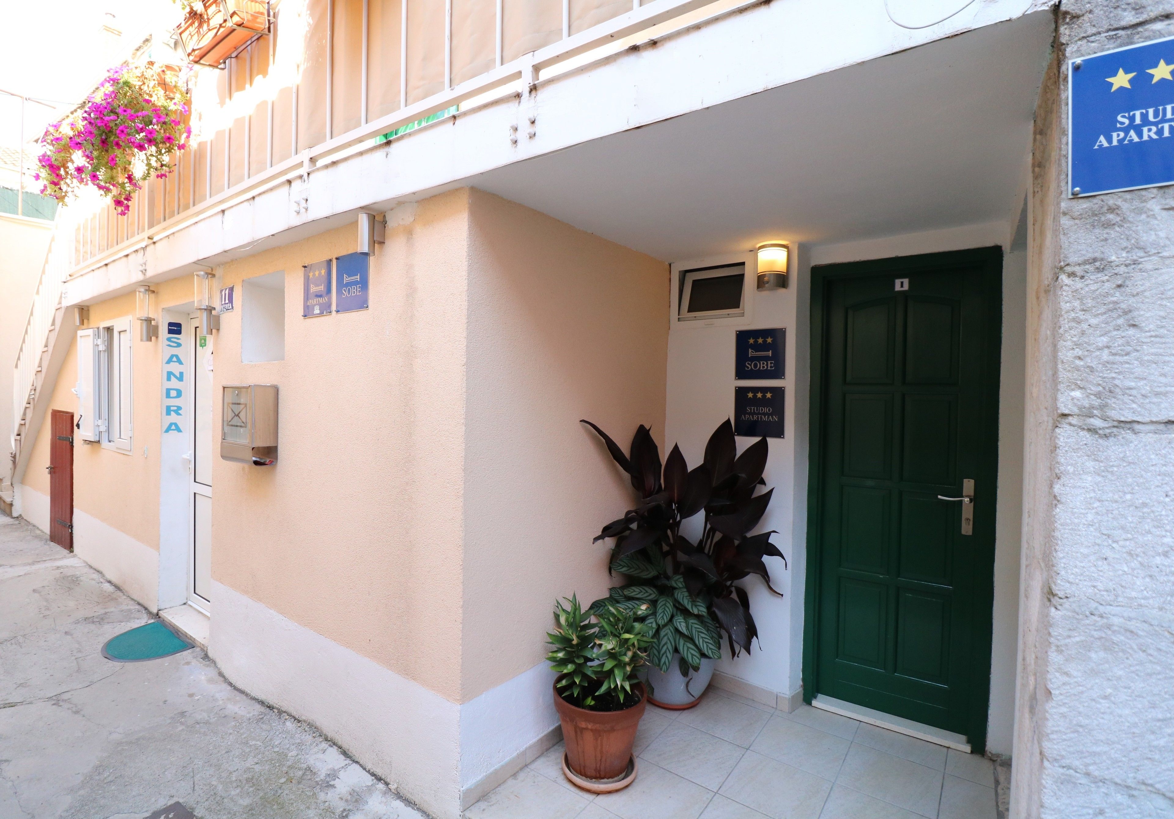 Un immeuble avec une porte verte, une enseigne indiquant « APPARTEMENTS-STUDIOS » et plusieurs plantes en pot.