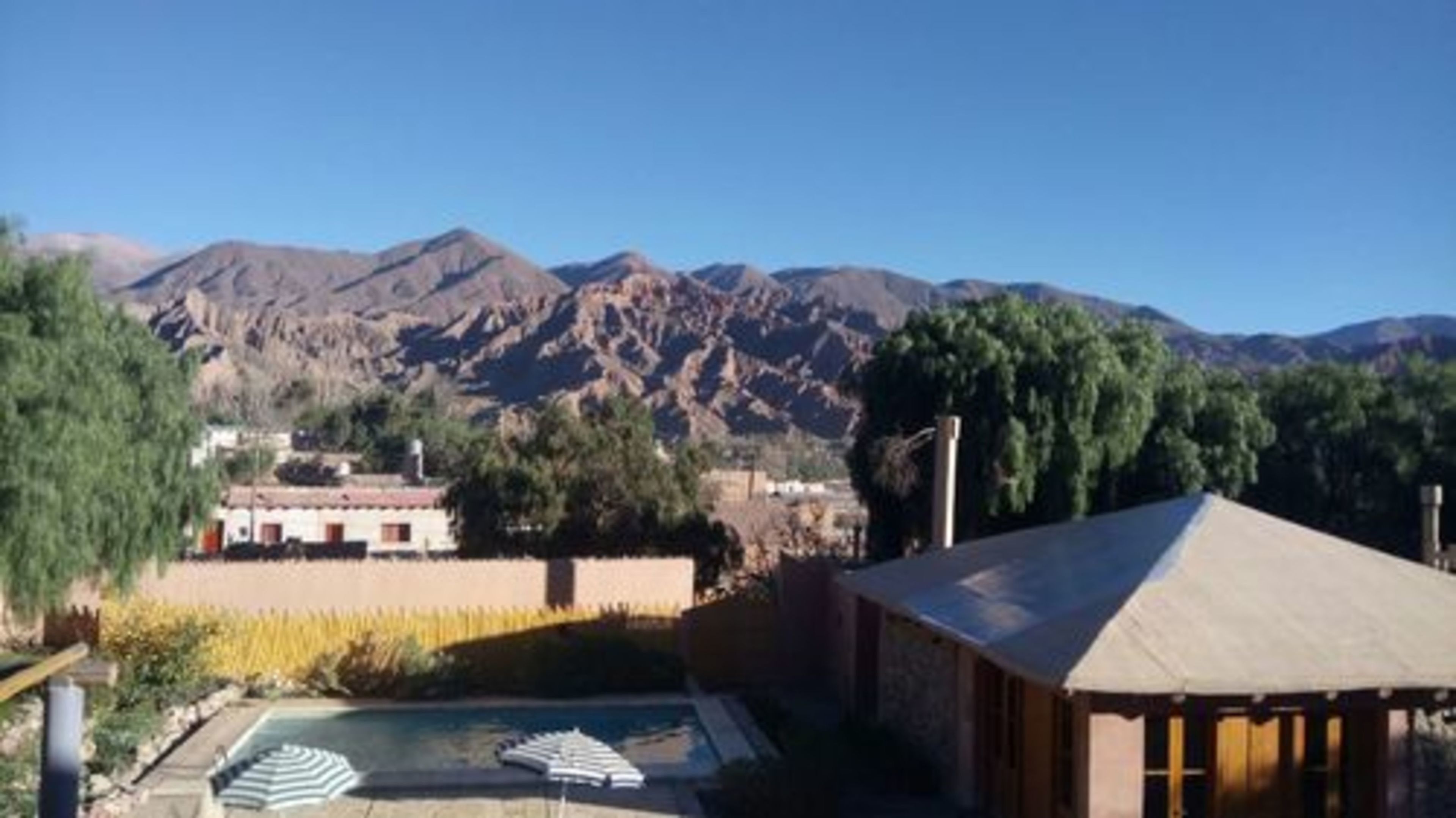 Zona de piscina con sillones de descanso, un edificio de techo rojo y montañas al fondo.