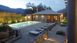 Zona de piscina con sillones reclinables, pared de piedra y un pabellón de madera.
