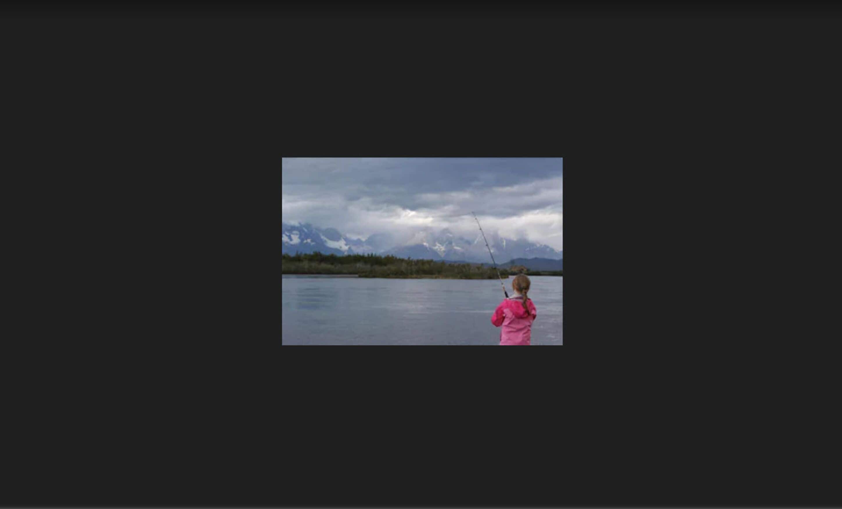 A person in a pink jacket fishing by a calm lake with mountains in the background.