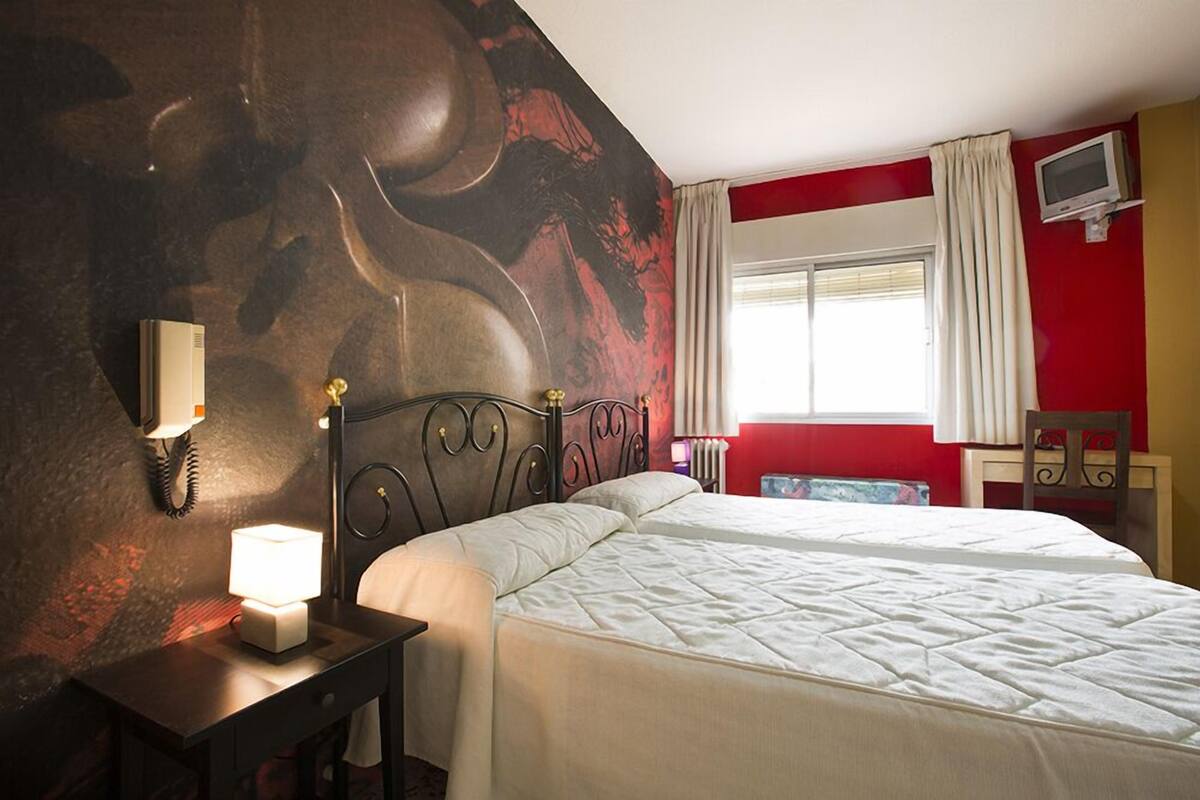 Una habitación de hotel con una cama grande, una mesita de noche con lámpara, un televisor en un mueble bajo y una ventana con cortinas.