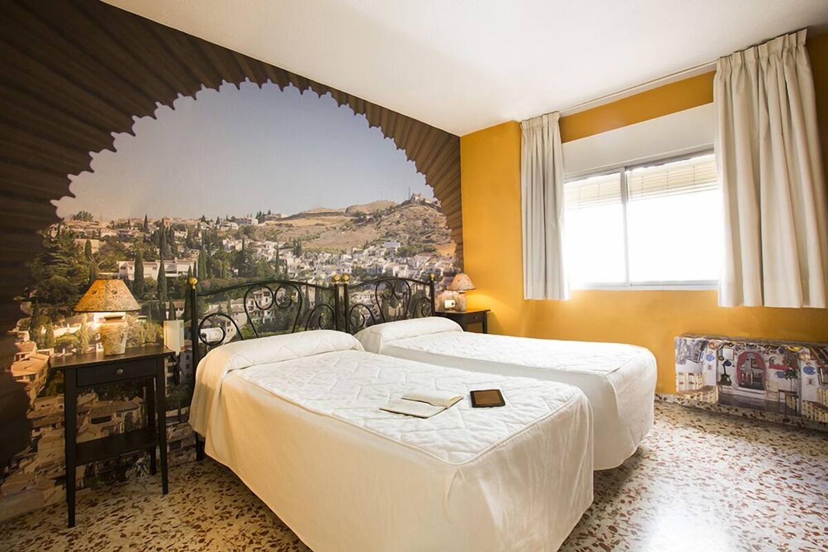 Habitación de hotel con dos camas, vista a un pueblo a través de un arco decorativo, una ventana con cortinas y una mesita con lámpara.
