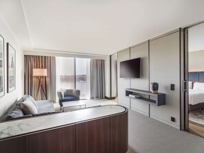 A modern hotel room with a large bed, a seating area with a sofa and armchair, a flat-screen TV mounted on the wall, and a view of a cityscape through large windows. - Radisson Blu Waterfront Hotel (Stockholm)