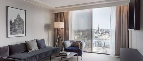 A modern hotel room with a large sofa, a smaller armchair, a coffee table, a floor lamp, and a large window offering a city view.