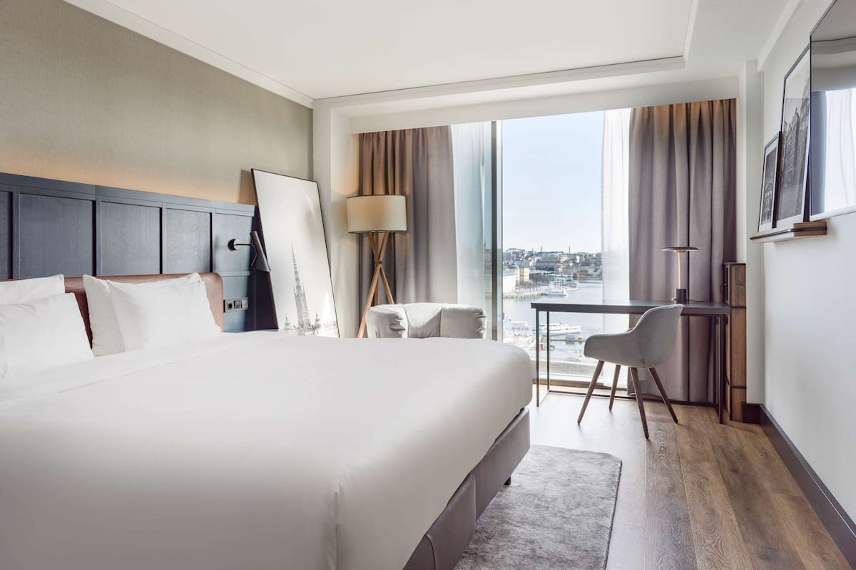 Una camera d'hotel con un letto grande, una scrivania, una sedia, una lampada e vista sulla città attraverso ampie finestre.