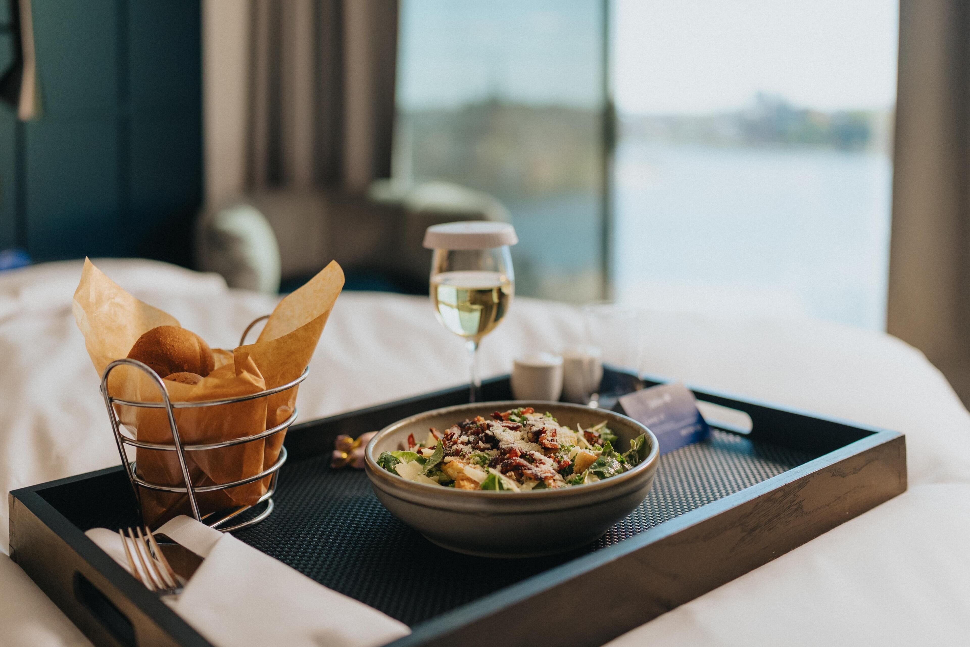 A tray with a bowl of salad, a glass of white wine, and a basket of bread rolls on a bed.