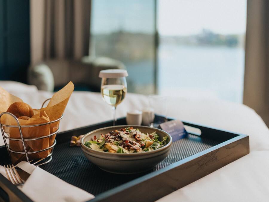 A tray with a bowl of salad, a glass of white wine, and a basket of bread rolls on a bed.