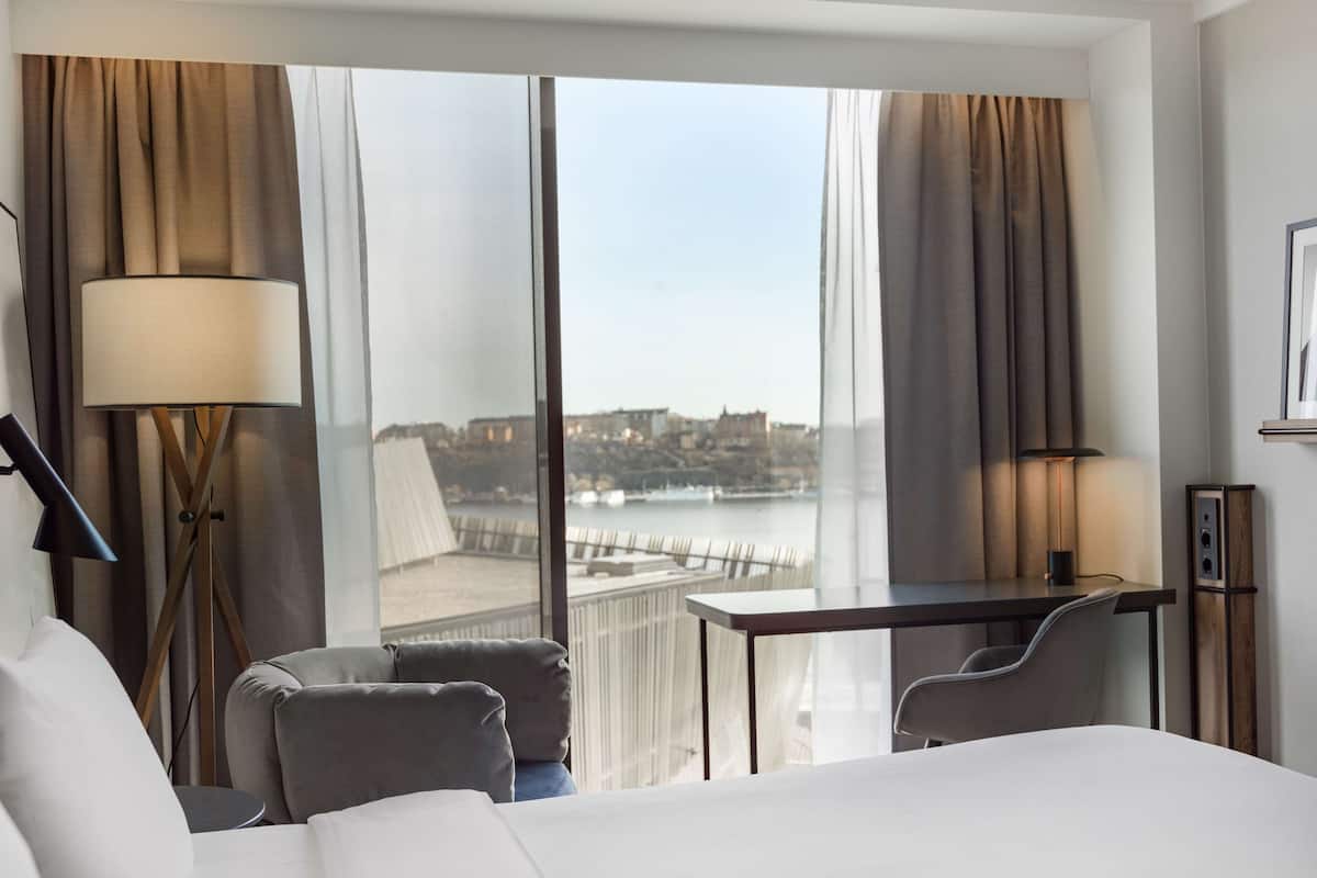 Camera d'albergo con un letto grande, due poltrone, un tavolino e vista su un corso d'acqua e degli edifici.