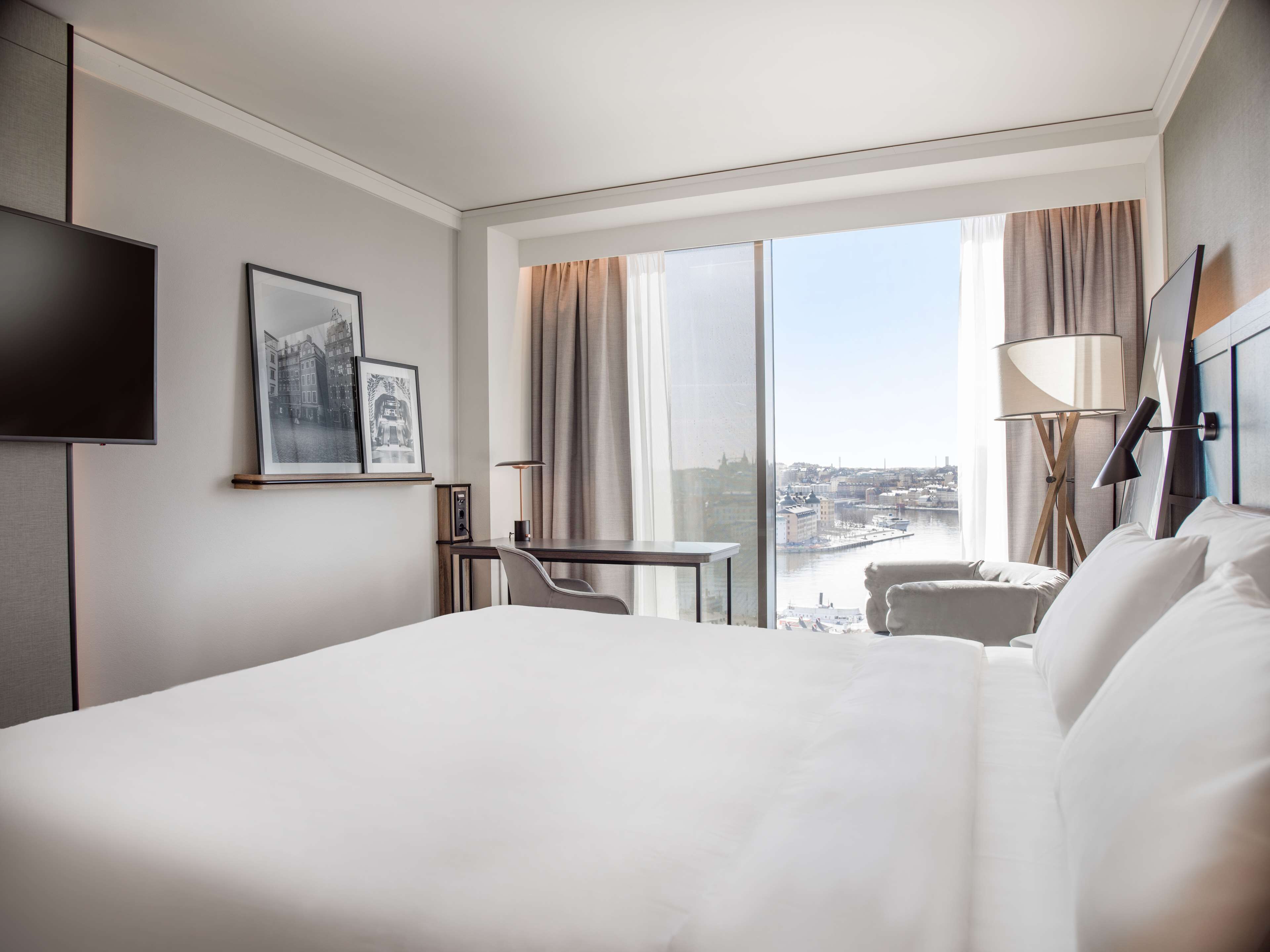Ein Hotelzimmer mit einem großen Bett, einem Schreibtisch, einem Stuhl, einem Fernseher und einem Fenster mit Blick auf die Stadt.