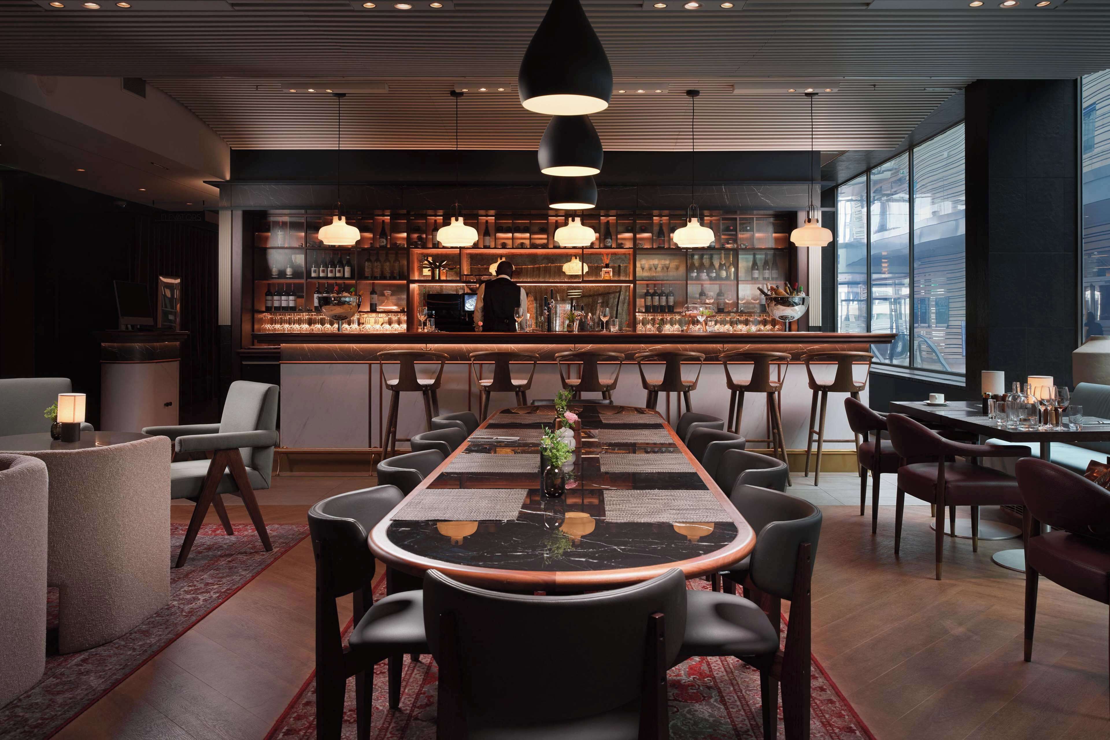 A modern bar area with a long table, multiple chairs, and a bartender.