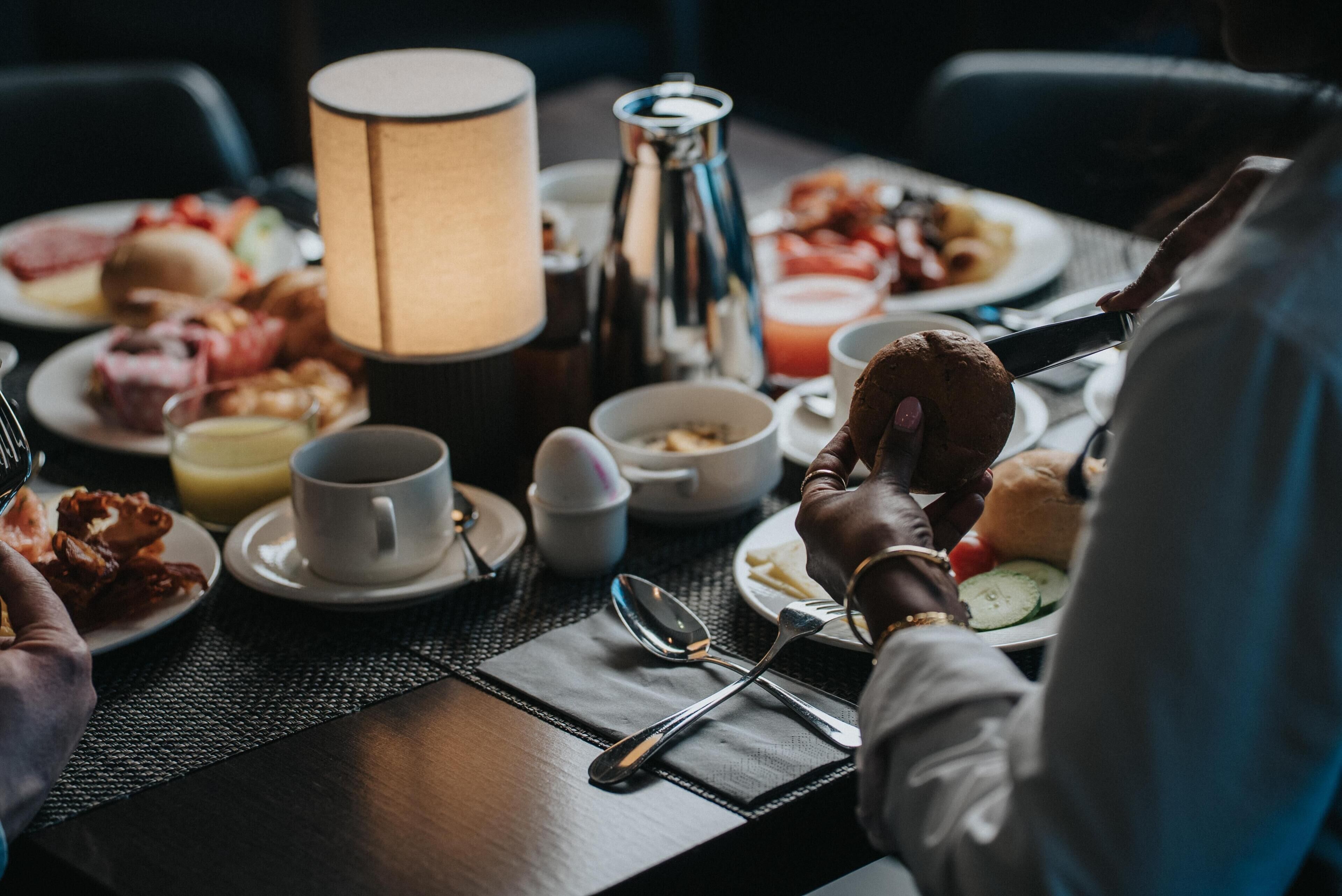 Mesa de café da manhã com pratos de comida, uma xícara de café, um abajur e uma pessoa comendo.