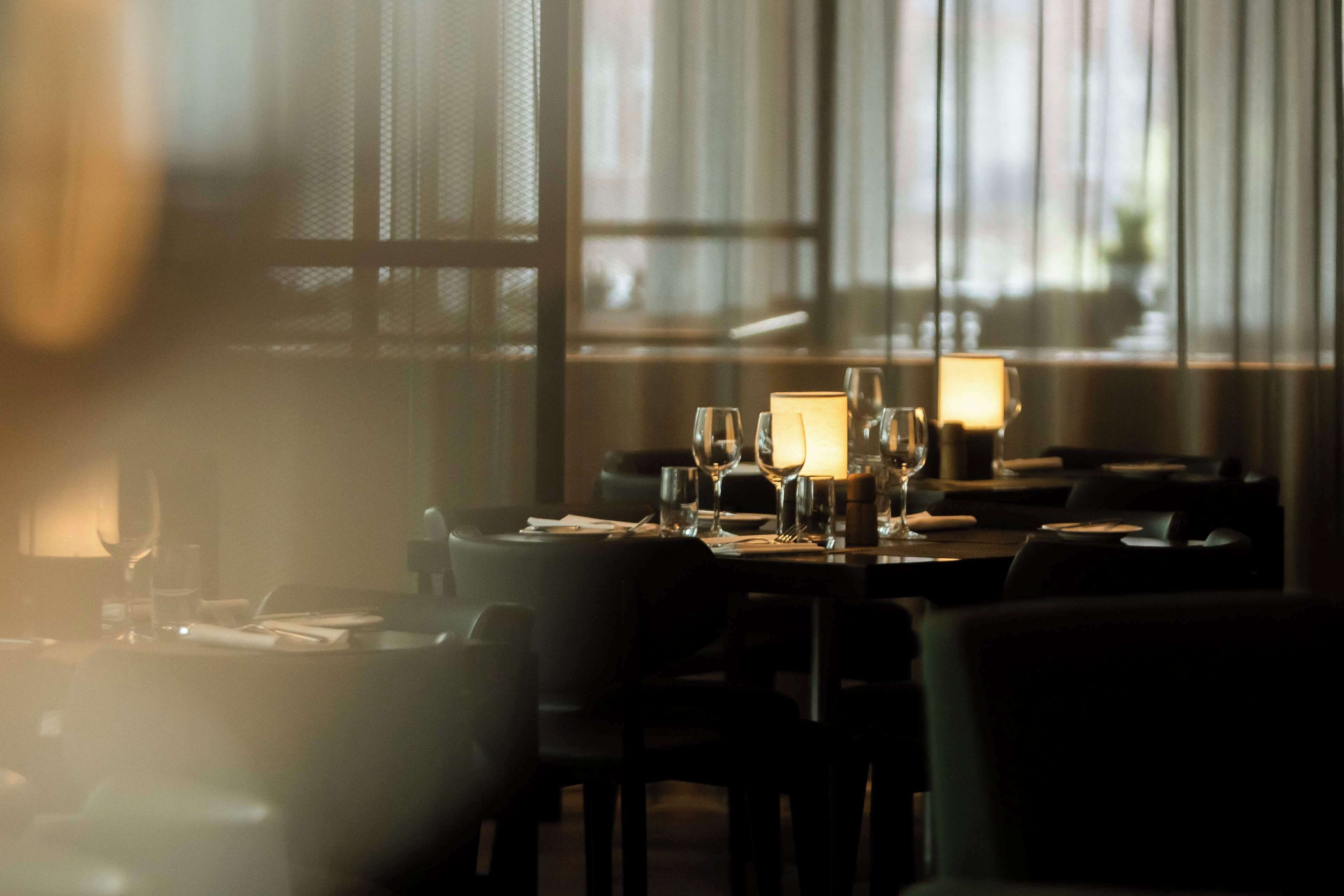 En restaurang med dämpad belysning, där borden är dukade för middag, och som präglas av en ombonad atmosfär och stora fönster.