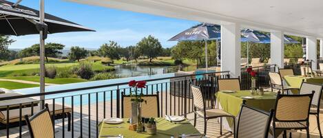 A covered outdoor dining area with tables and chairs, a pool, and a view of a golf course and lake.