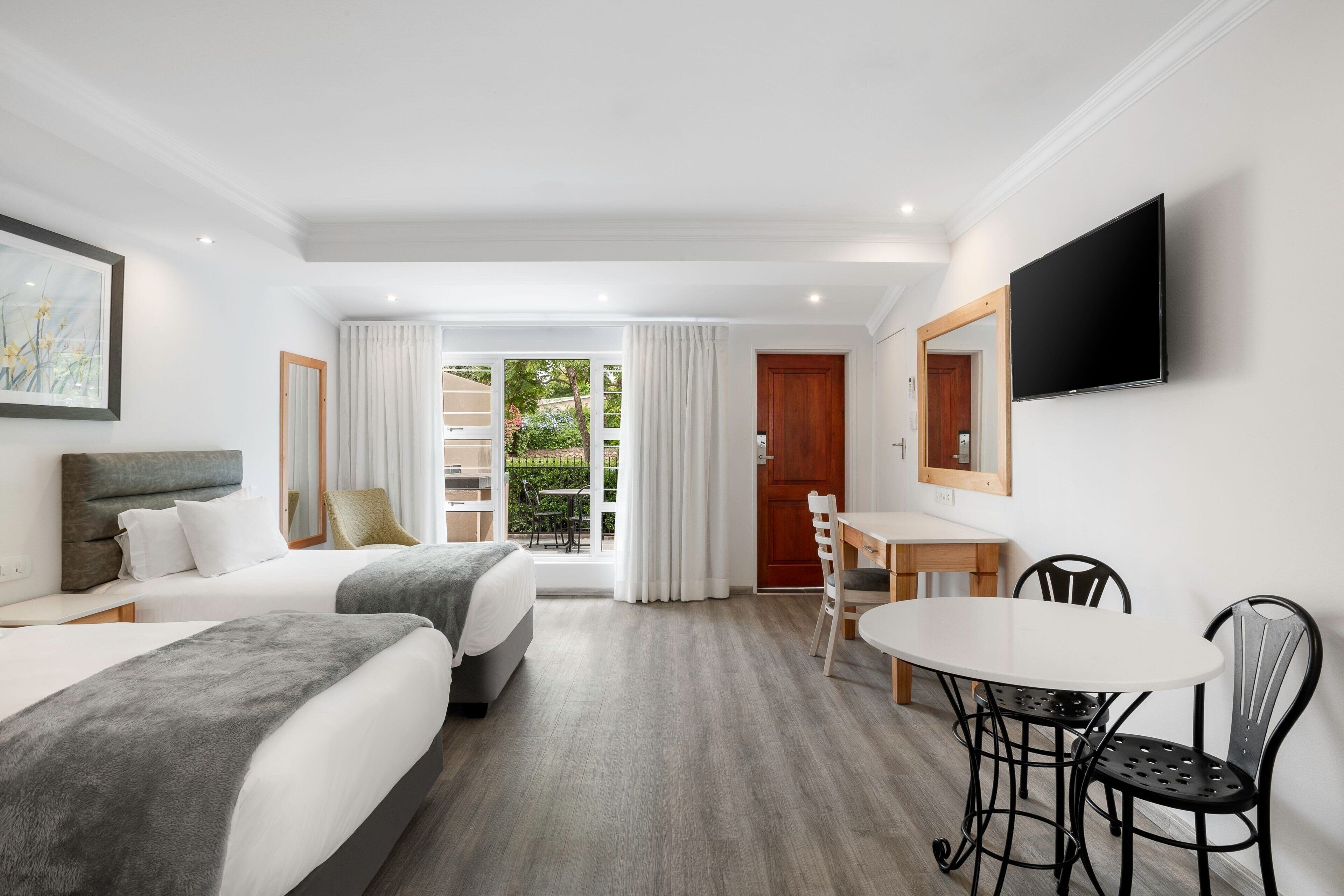 A hotel room with two beds, a dining table with chairs, a TV mounted on the wall, and a view of the outdoors through a glass door.