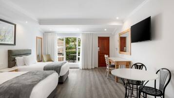 A hotel room with two beds, a dining table with chairs, a TV mounted on the wall, and a view of the outdoors through a glass door.