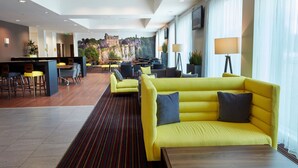 Un moderno vestíbulo de hotel con una zona de estar central con sofás amarillos, una zona de bar con taburetes y un gran ventanal con vistas a un edificio histórico.