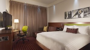 Habitación de hotel con cama, escritorio con televisor, una silla y un amplio ventanal con cortinas.