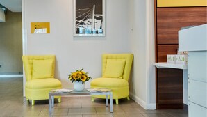 Un área de recepción con dos sillones amarillos, una mesita con un jarrón de girasoles y un cuadro en la pared.