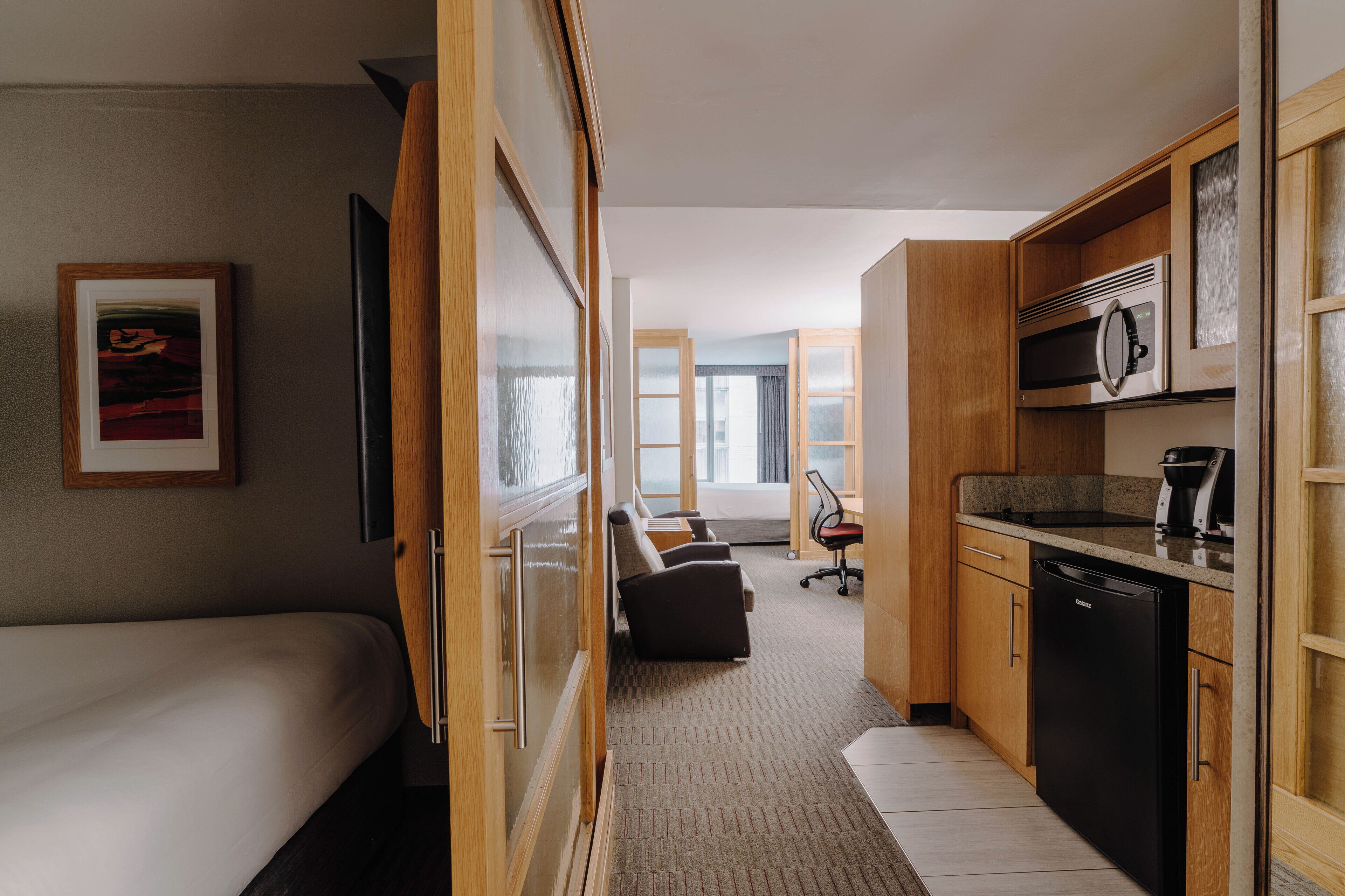 A compact hotel room with a bed, a small kitchenette, a seating area with a sofa, and a desk.