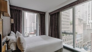 A hotel room with a large bed, a desk, and a chair. There are two windows with curtains, and a view of tall buildings outside. - The Jewel Hotel, New York (New York)