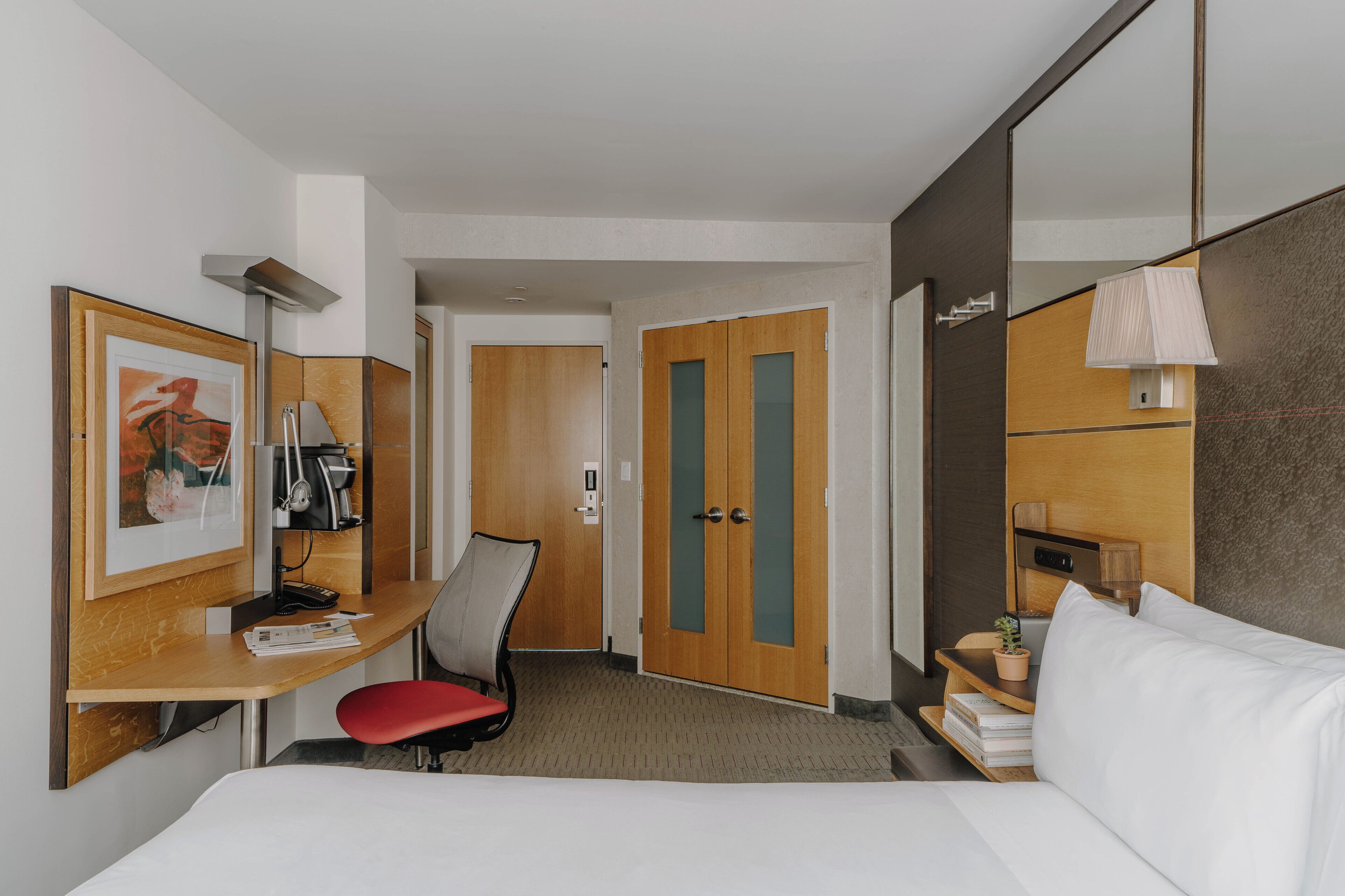 Une chambre d’hôtel comprenant un lit, un bureau avec une chaise, un miroir, une porte et une petite table avec une lampe et une plante.