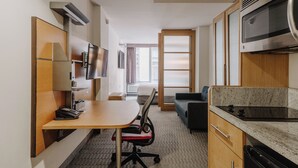 A compact hotel room with a kitchenette, a desk with a chair, a sofa, and a TV mounted on the wall. - The Jewel Hotel, New York (New York)