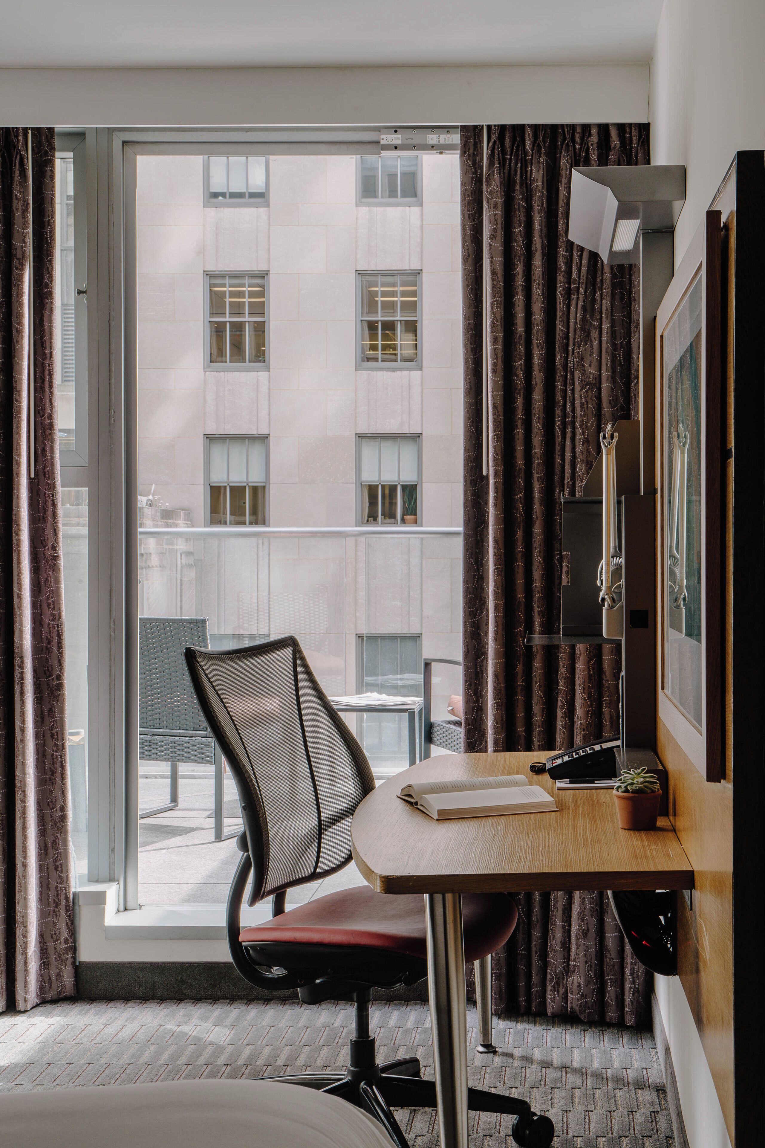 Habitación de hotel con un ventanal grande, un escritorio con silla, una planta en maceta y un cuadro en la pared.