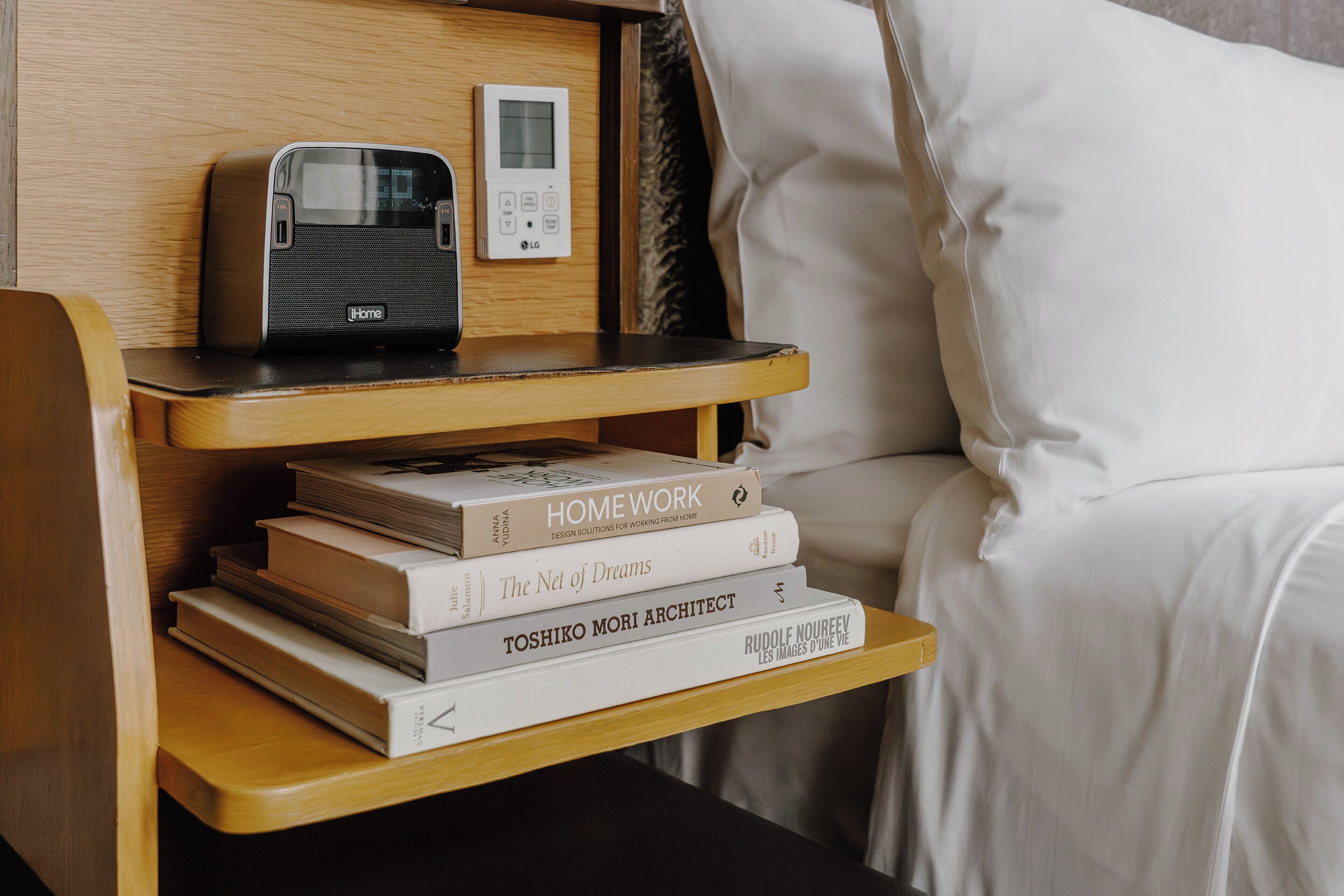 A bedside table with a speaker, a stack of books, and a digital thermostat.