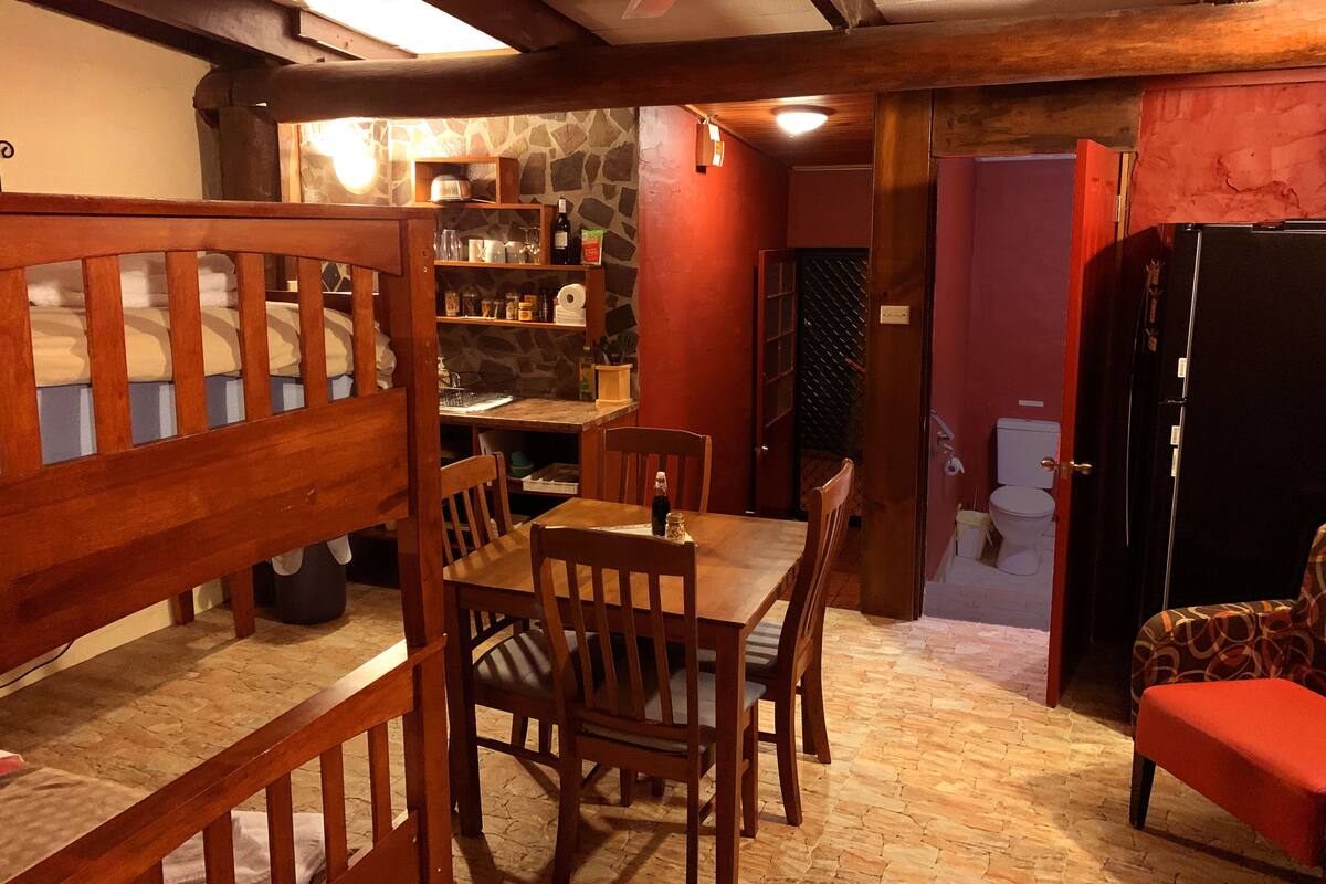 A cozy room with a wooden dining table, chairs, a bench, a refrigerator, and a kitchen area with shelves and a stone wall.
