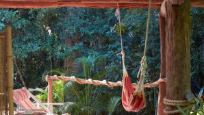 Una hamaca colgada entre dos árboles en un espacio exterior techado, con un banco de madera y una mesa.