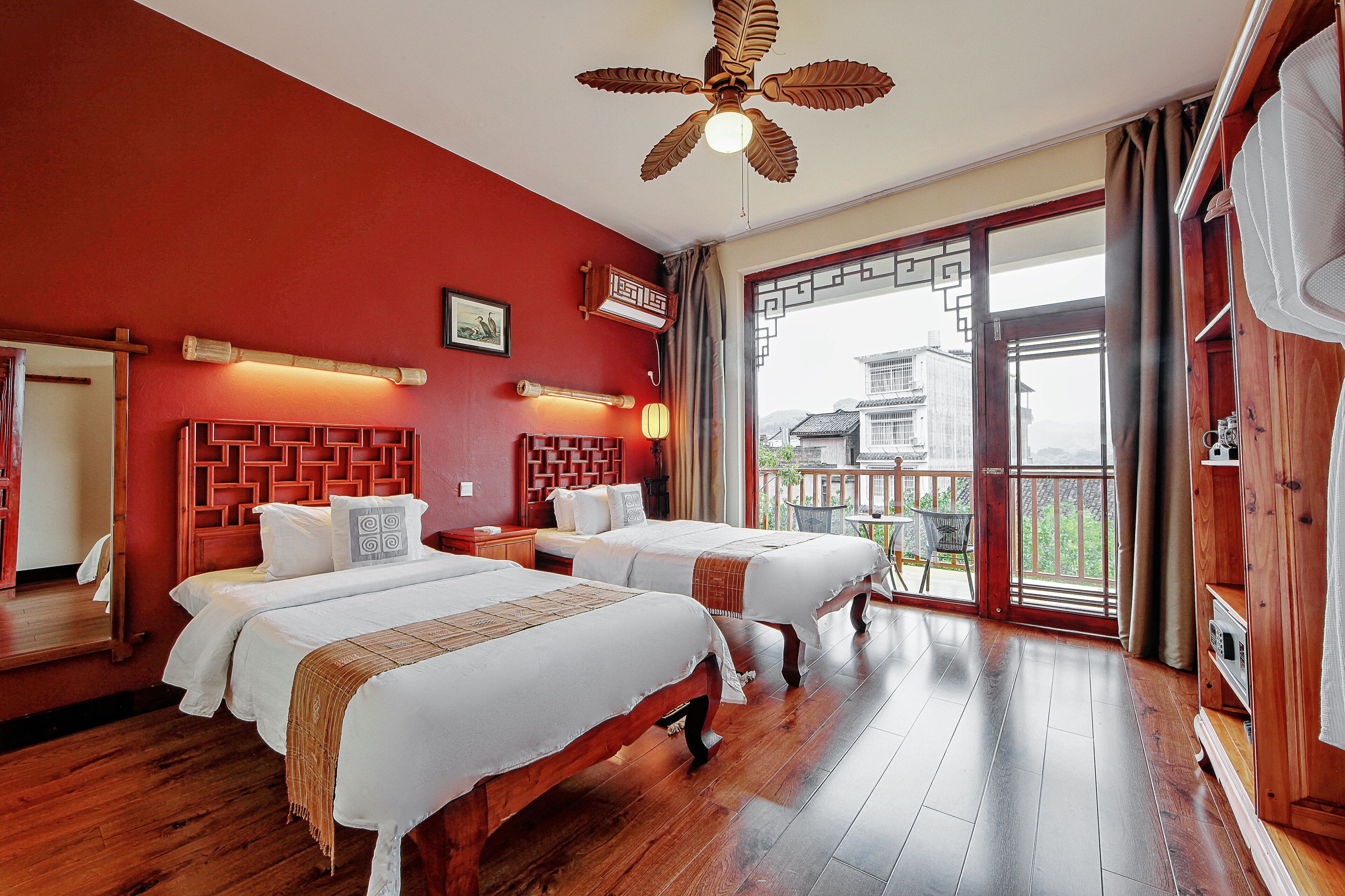 A hotel room with two beds, wooden flooring, a ceiling fan, and a balcony with a view.