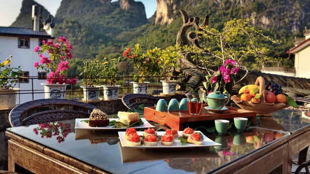 Una mesa servida con comida y bebidas, rodeada de plantas en macetas y un paisaje montañoso de fondo.