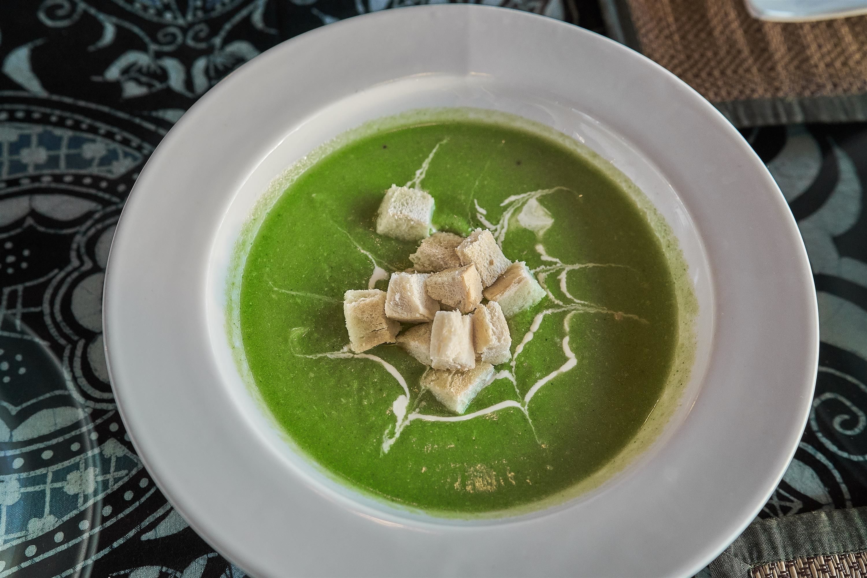 A bowl of green soup with croutons on a patterned tablecloth.